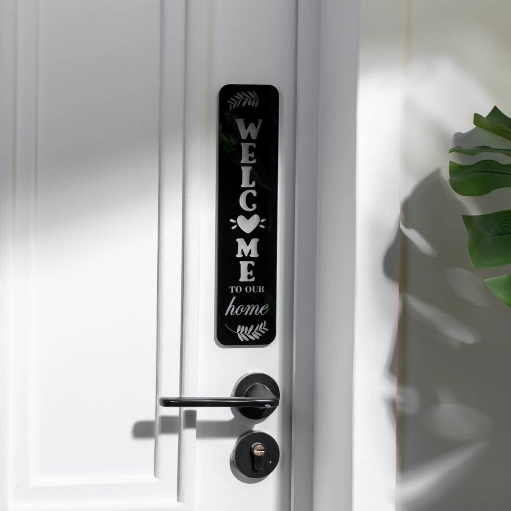 Placa vertical de acrilico negro grabada con la palabra welcome y un corazon, colgada junto al picaporte de una puerta blanca