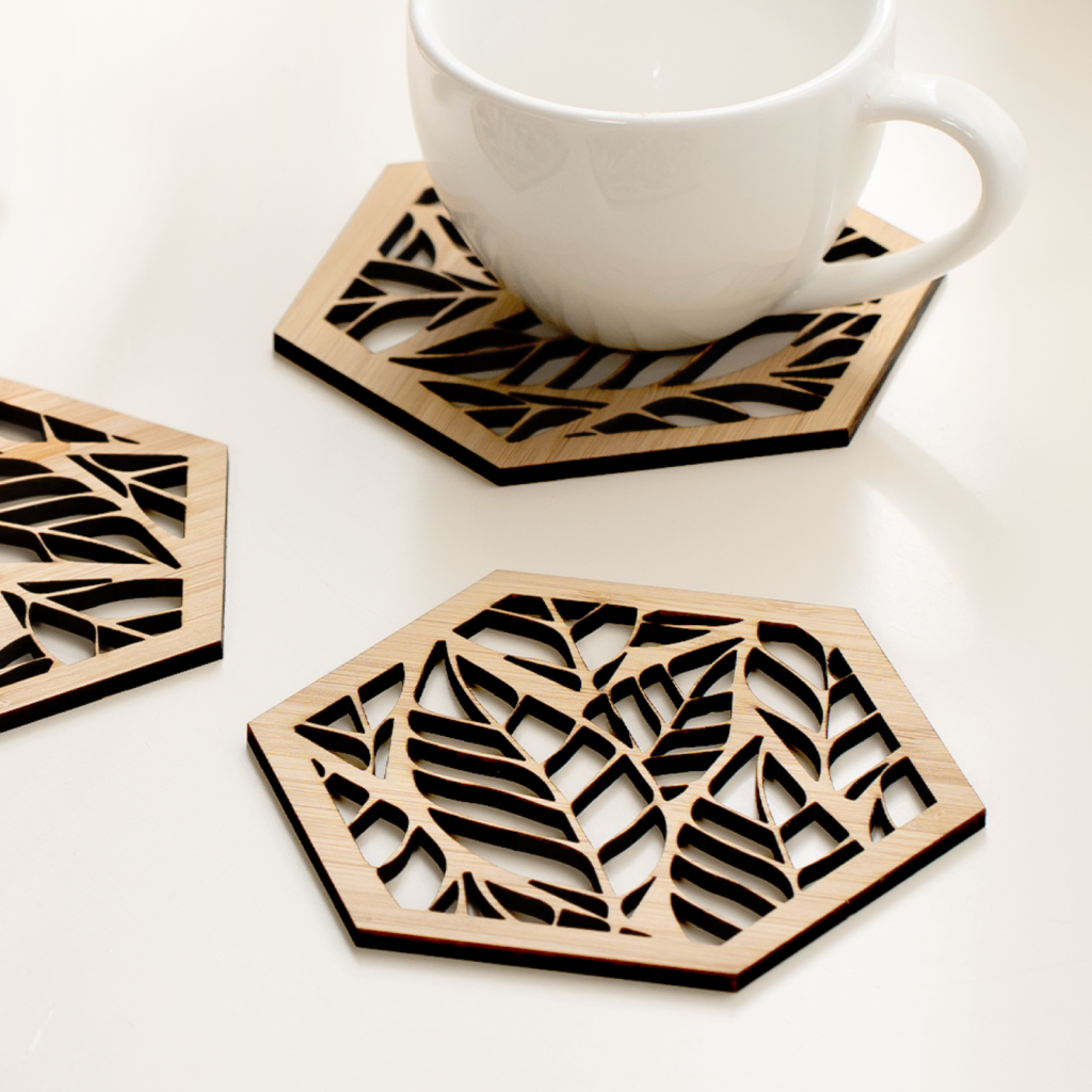 Posavasos hexagonales calados en tablero de bambu, con un motivo de hojas, junto a una taza de cafe
